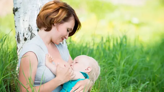 Frau stillt Baby in Graswiese mit einem Baum dahinter