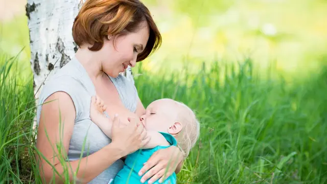 Frau stillt Baby in Graswiese mit einem Baum dahinter