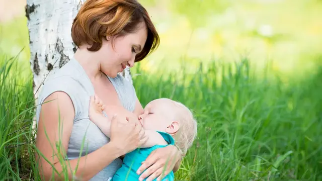 Frau stillt Baby in Graswiese mit einem Baum dahinter