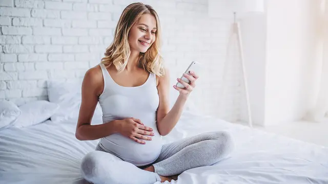 Schwangere Frau sitzt mit Handy in der Hand auf dem Bett