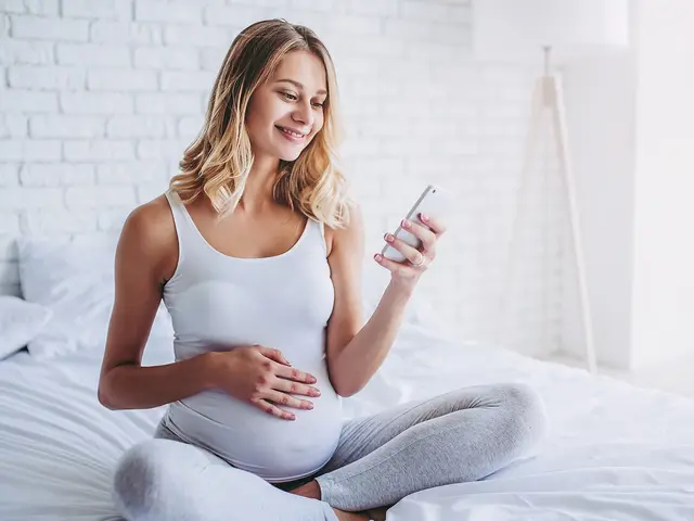 Schwangere Frau sitzt mit Handy in der Hand auf dem Bett